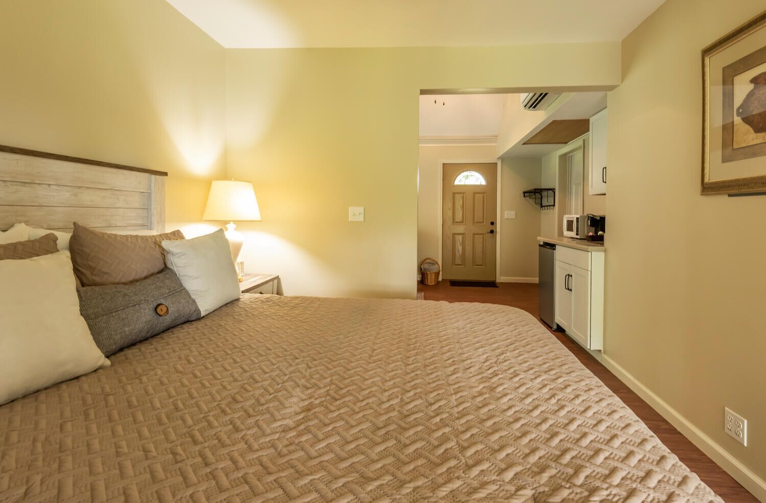 A hotel room with a large bed, a bedside lamp, a door leading to a kitchen, and a framed picture on the wall.
