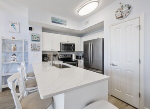 A modern kitchen with white cabinetry, a stainless steel refrigerator, a microwave, and a sink. There are decorative items on the shelves and a clock on the wall.