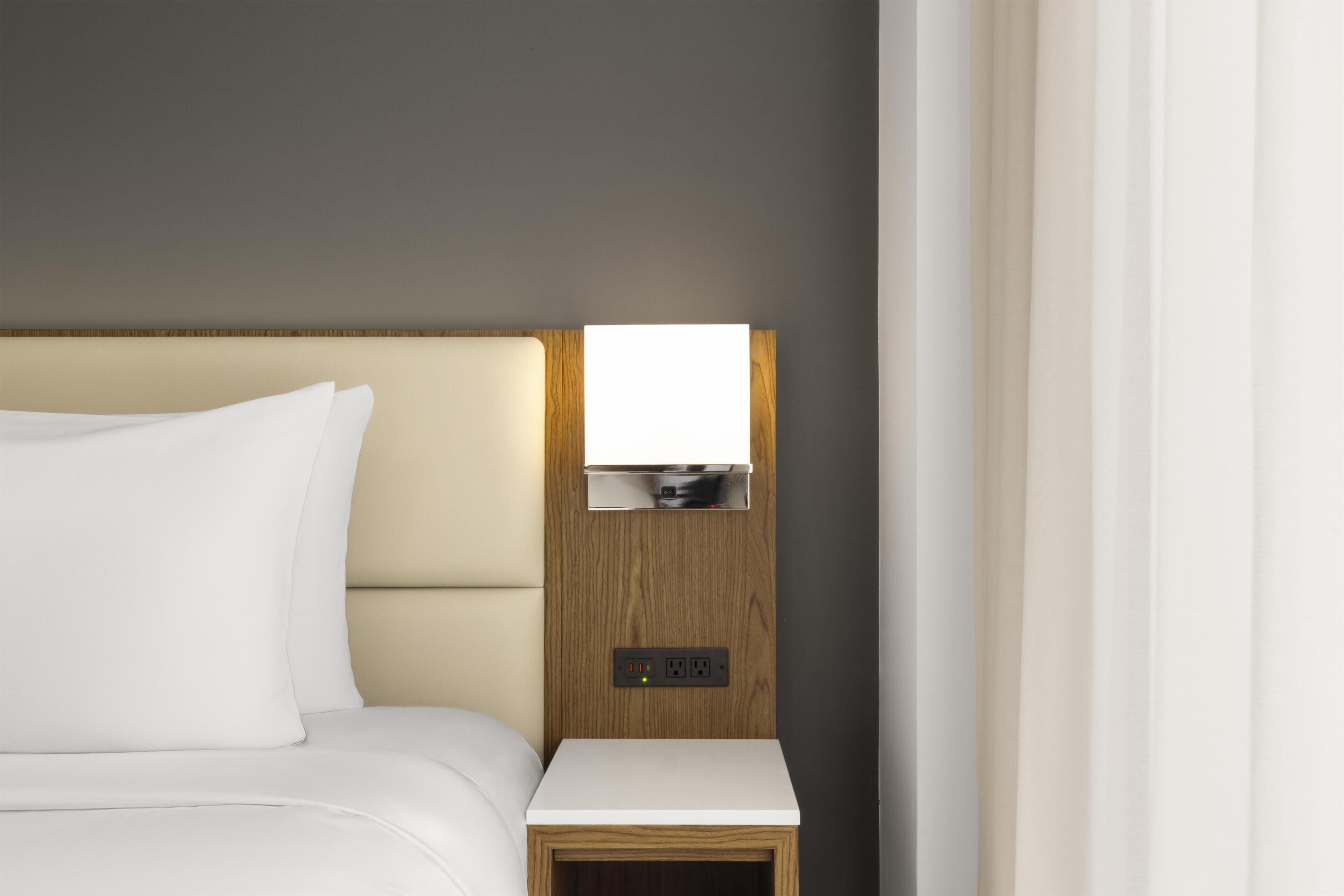 Een hotelkamer met een bed, een nachtkastje met een lamp en een wandcontactdoos.