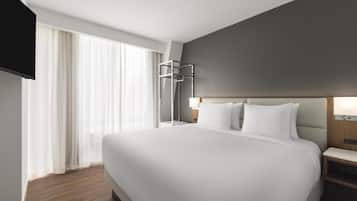A modern hotel room with a large bed, a TV mounted on the wall, and a window with sheer curtains.