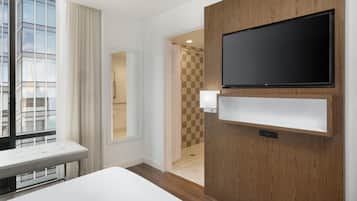Une chambre d’hôtel dotée d’une grande fenêtre, d’un téléviseur à écran plat fixé sur un panneau en bois, d’une table de chevet et d’un espace douche visible à travers une porte ouverte.