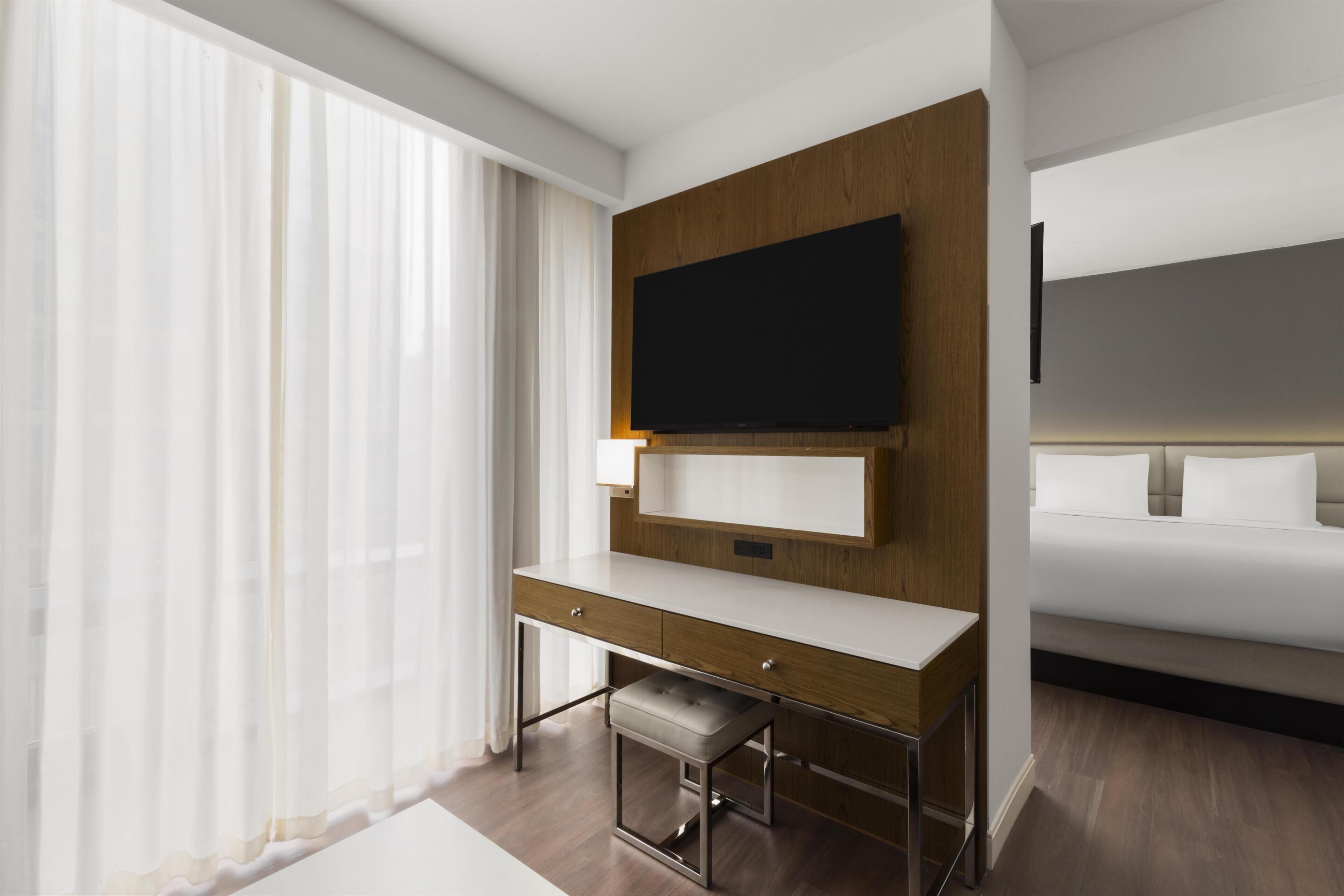 Quarto de hotel moderno com uma cama grande, uma escrivaninha com cadeira, televisão na parede e cortinas finas na janela.
