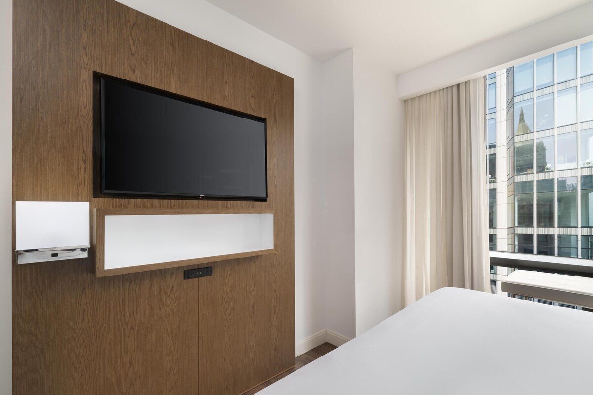 Une chambre d’hôtel équipée d’un téléviseur à écran plat fixé au mur, d’une étagère intégrée, d’une grande fenêtre avec des rideaux et offrant une vue sur un bâtiment doté d’une flèche.