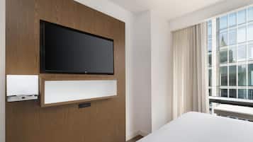 Habitación de hotel con televisión de pantalla plana montada, estante empotrado, ventana grande con cortinas y vista a un edificio con aguja.