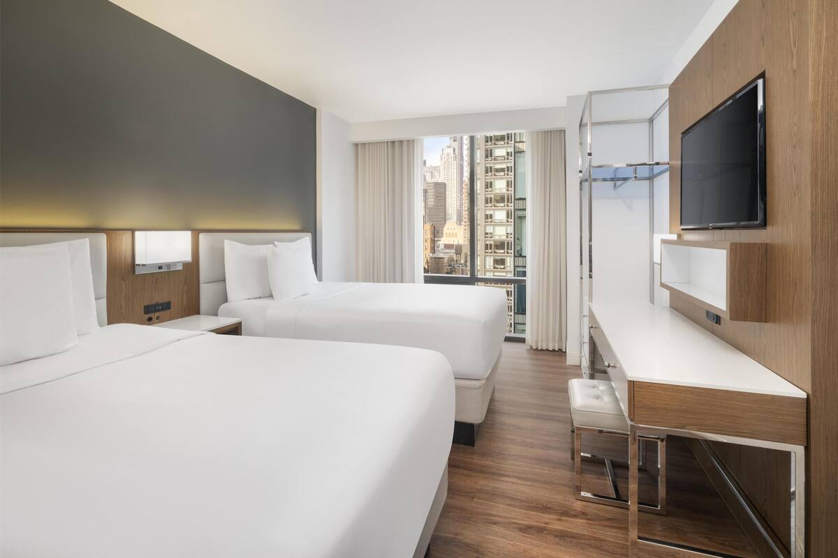 Ein Hotelzimmer mit zwei Betten, einem Schreibtisch, einem Fernseher und einem großen Fenster mit Blick auf die Stadt.