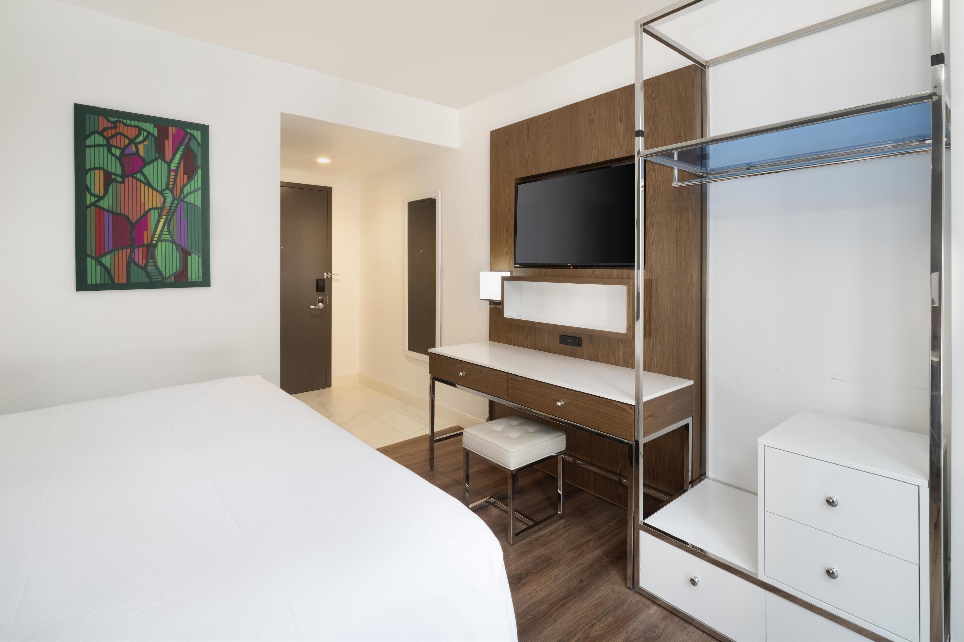 Una habitación de hotel moderna con una cama, un escritorio con silla, un televisor montado en un panel de madera y un armario con espejo.