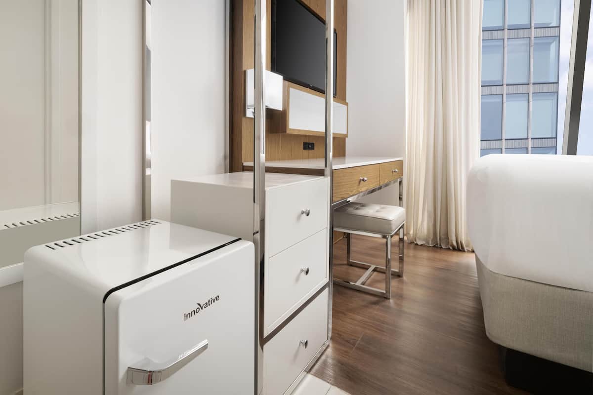 Ein Hotelzimmer mit einer kompakten Küchenzeile, einem Schreibtisch mit Stuhl, einem Bett und einem großen Fenster mit Vorhängen.