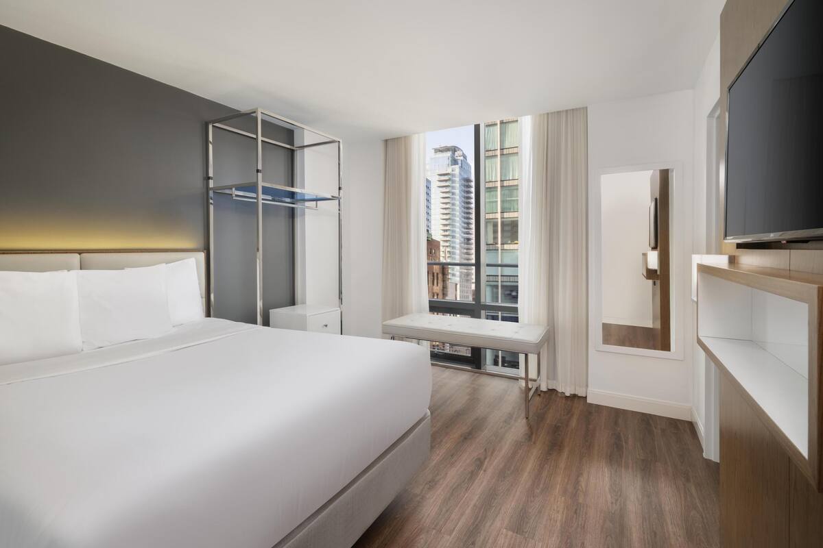 Ein modernes Hotelzimmer mit einem großen Bett, einem Fernseher, einem Schreibtisch und einem Fenster mit Blick auf die Stadt.