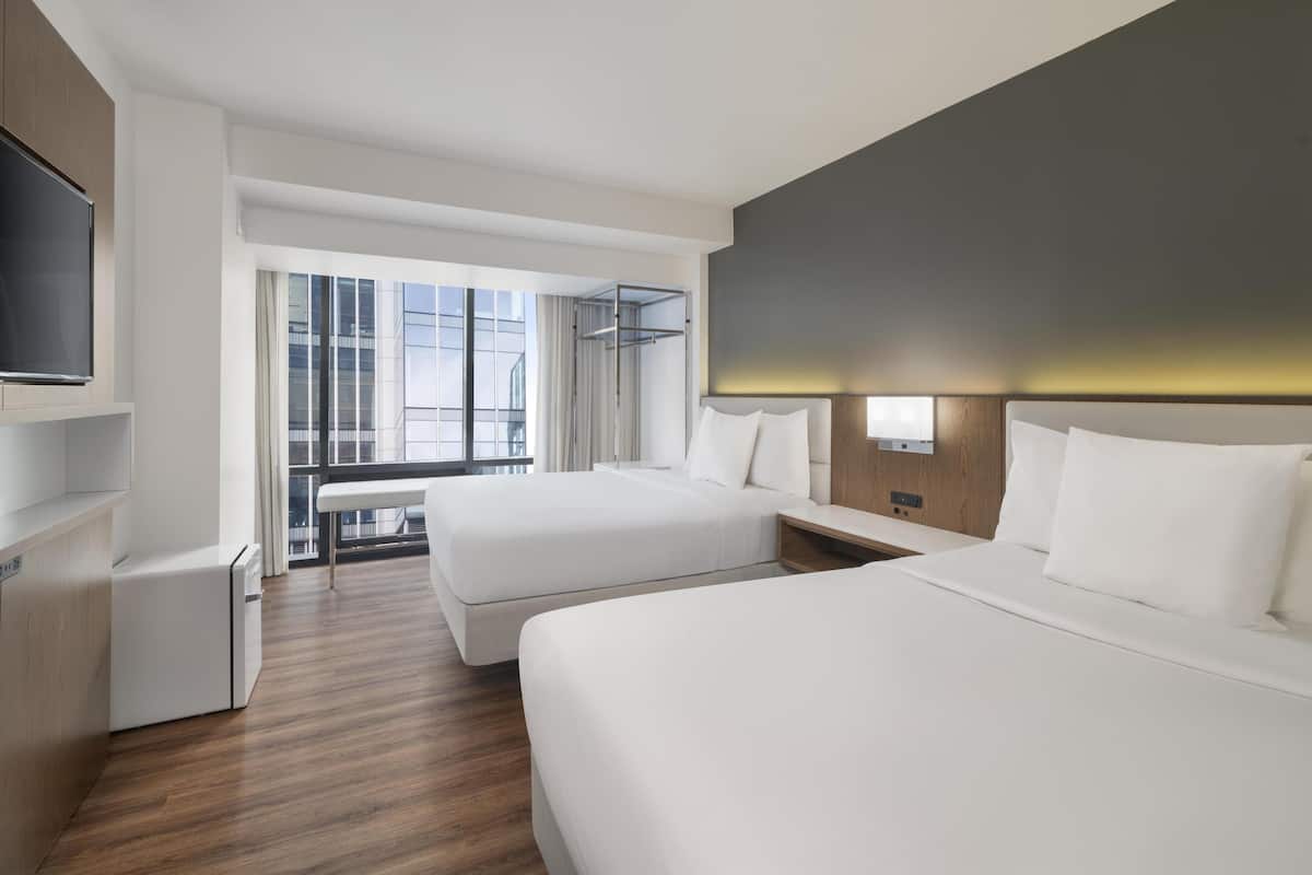 Ein Hotelzimmer mit zwei Betten, einem Fernseher, einem Schreibtisch und einem großen Fenster mit Blick auf Gebäude.
