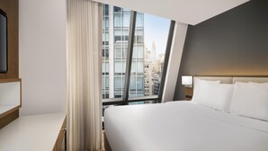 Een moderne hotelkamer met een groot bed, een bureau en uitzicht op een stadsgezicht door middel van kamerhoge ramen.
