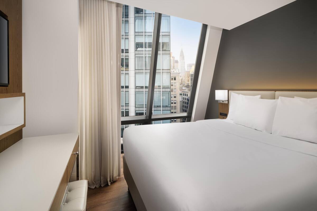 Ein modernes Hotelzimmer mit einem großen Bett, einem Schreibtisch und Blick auf eine Stadtlandschaft durch raumhohe Fenster.