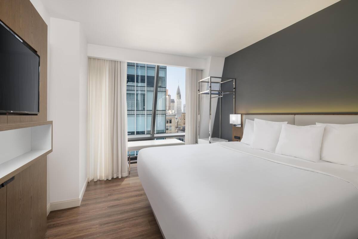 Ein modernes Hotelzimmer mit einem großen Bett, einem Flachbildfernseher, einem Schreibtisch und Blick auf die Skyline der Stadt.