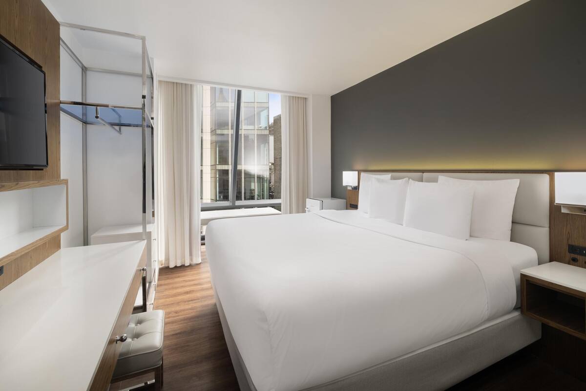 Ein modernes Hotelzimmer mit einem großen Bett, einem Schreibtisch, einem Fernseher und Blick auf die Stadt durch große Fenster.