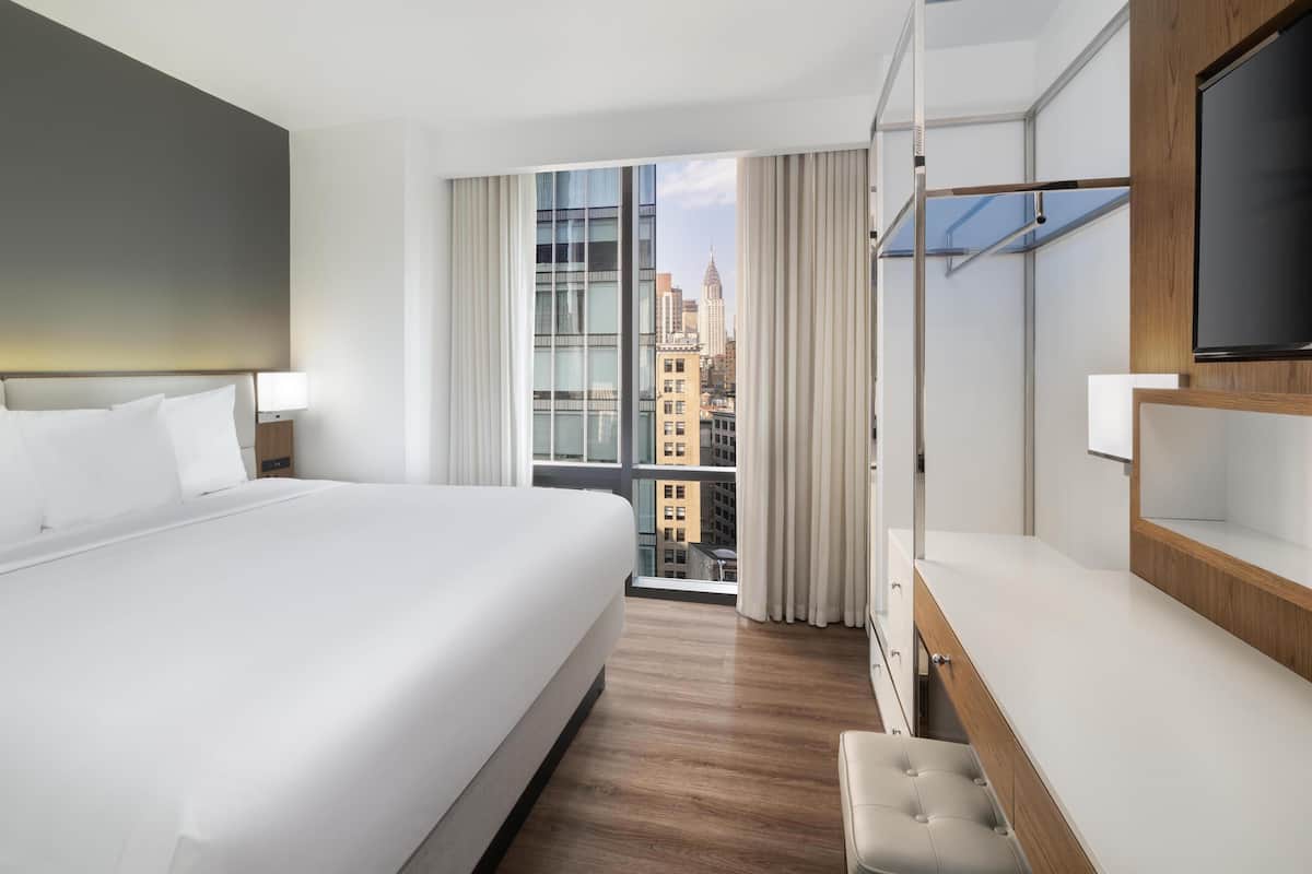 Ein Hotelzimmer mit einem großen Bett, einem Fernseher, einem Balkon mit Vorhängen und Blick auf die Stadt.