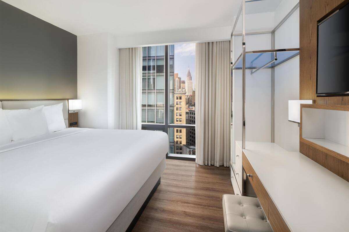 Ein Hotelzimmer mit einem großen Bett, einem Fernseher, einem Schreibtisch und Blick auf die Stadt.