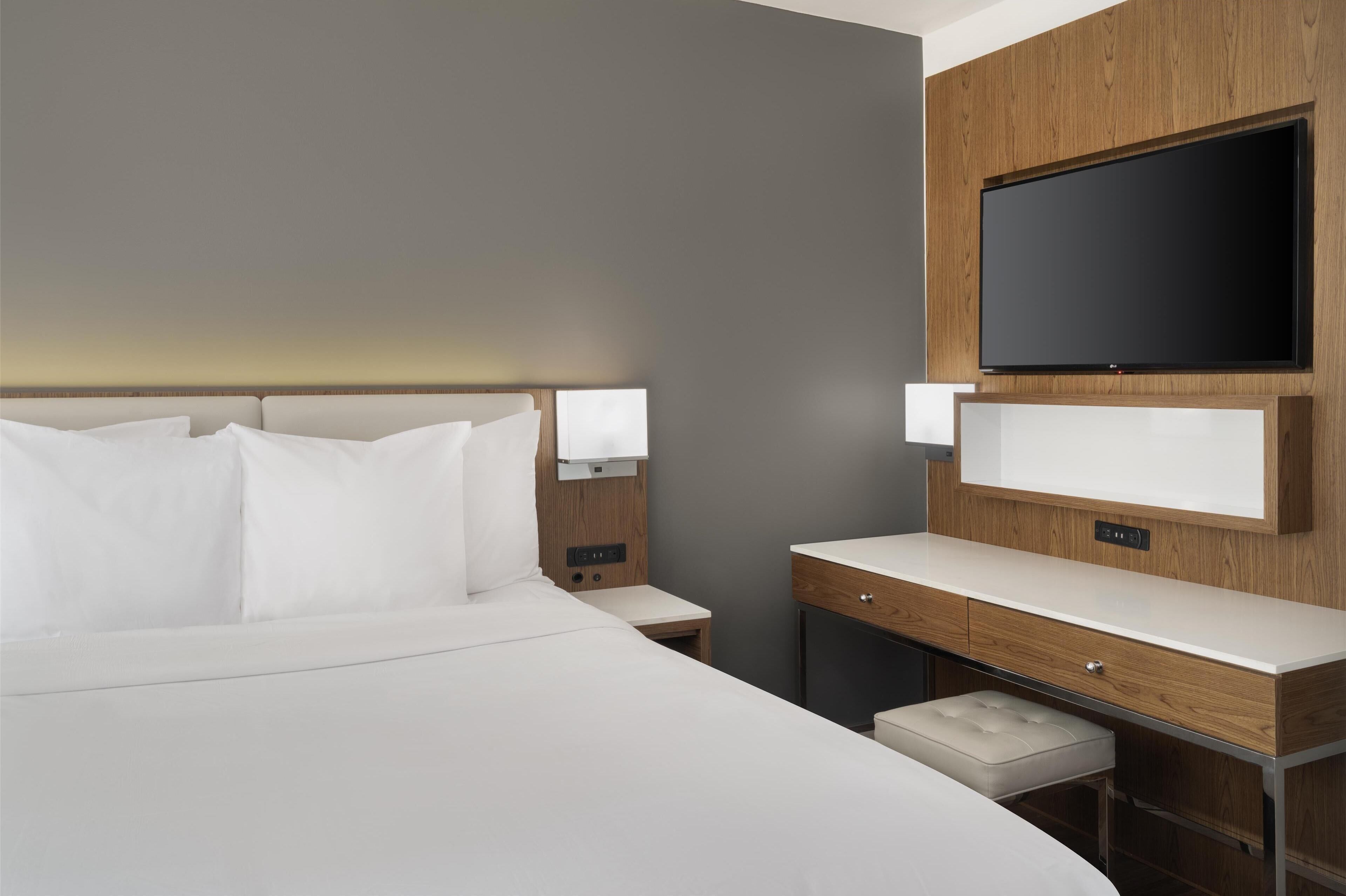 A modern hotel room with a large bed, a bedside table, a desk with a chair, and a flat-screen TV mounted on the wall.