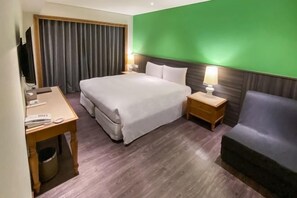 Habitación de hotel con cama, escritorio, silla, televisor y una pared verde decorativa.