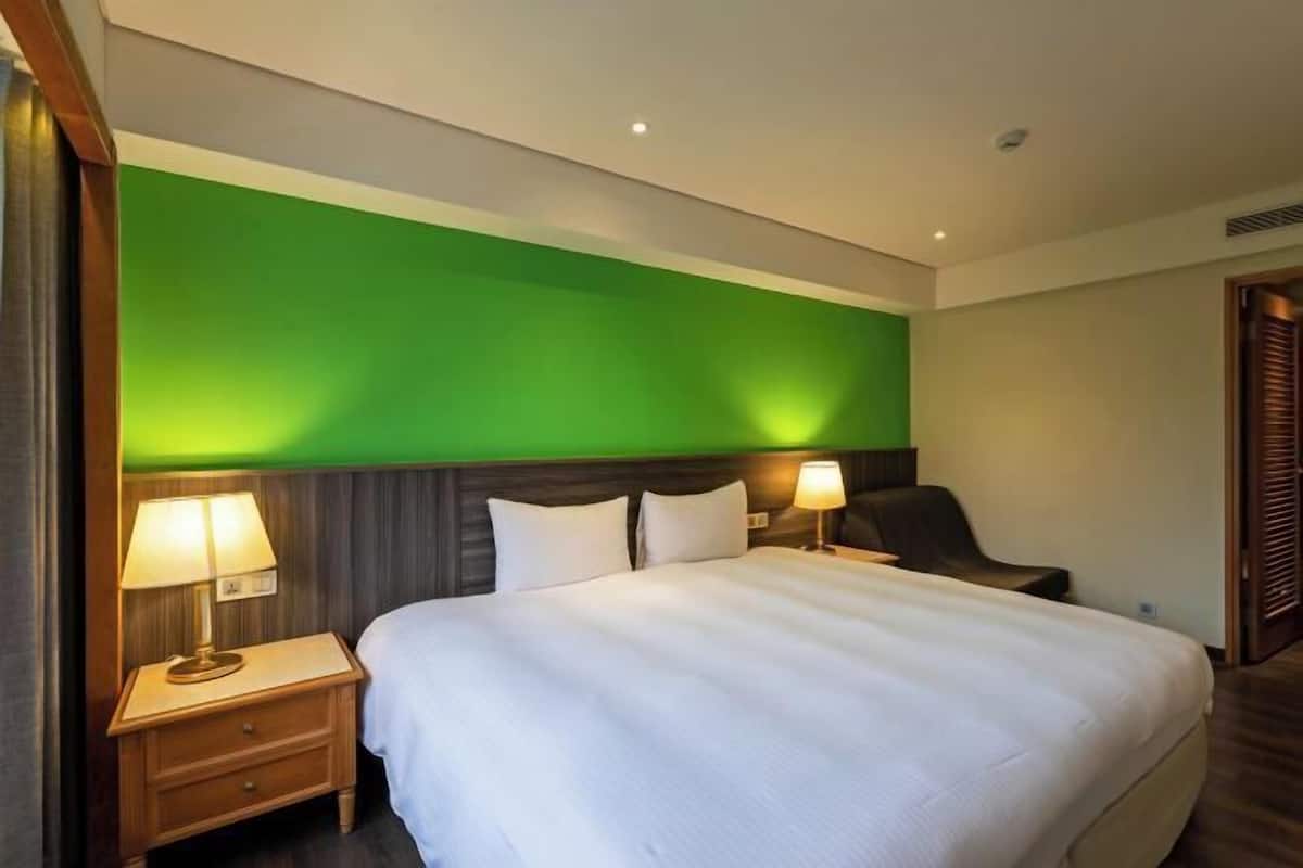A hotel room with a large bed, two bedside tables with lamps, a dark headboard, and a green accent wall.