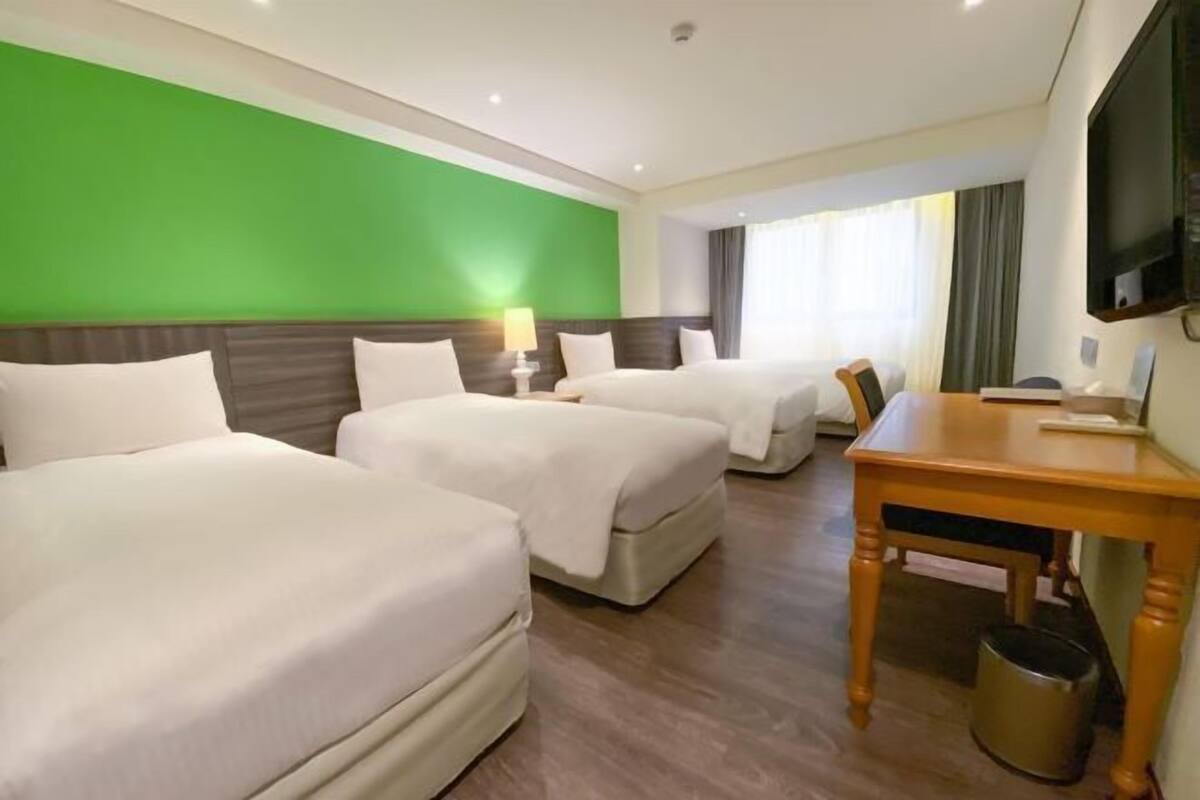 A hotel room with two beds, a wooden table, a TV, and a green accent wall.