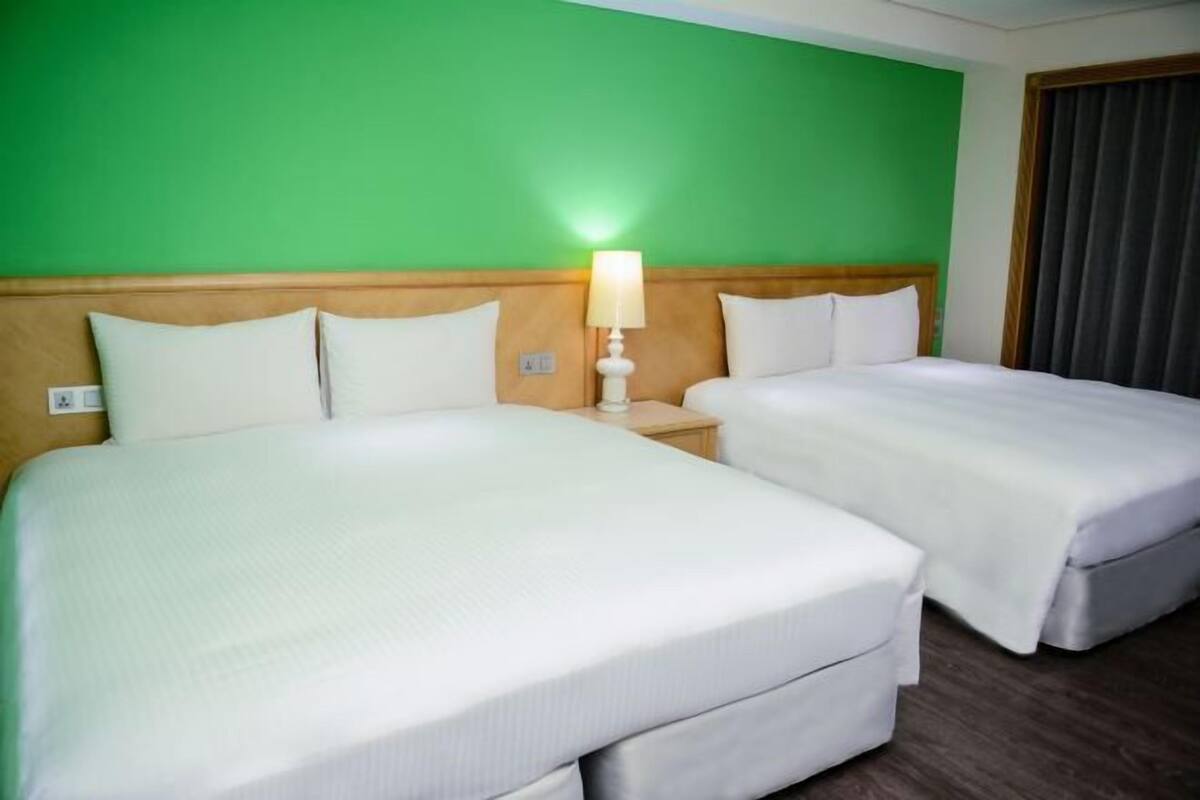 A hotel room with two beds, a nightstand with a lamp, and a green wall.