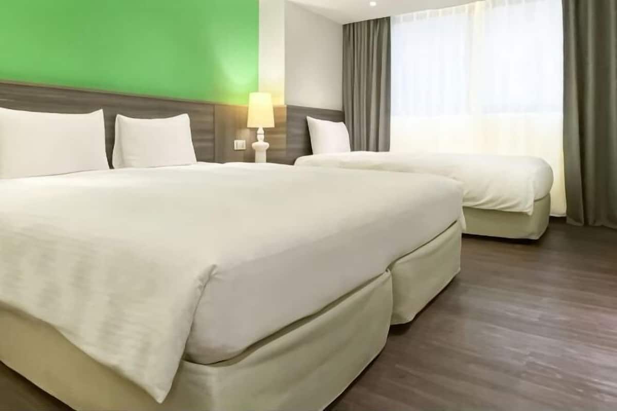 A hotel room with two beds, a nightstand with a lamp, a green accent wall, and a window with curtains.