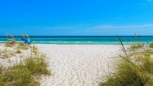 Ein Strand mit blauem Himmel, türkissem Meer und weißem Sand. Einige Gräser und wenige Strandsonnenstühle sind zu sehen.
