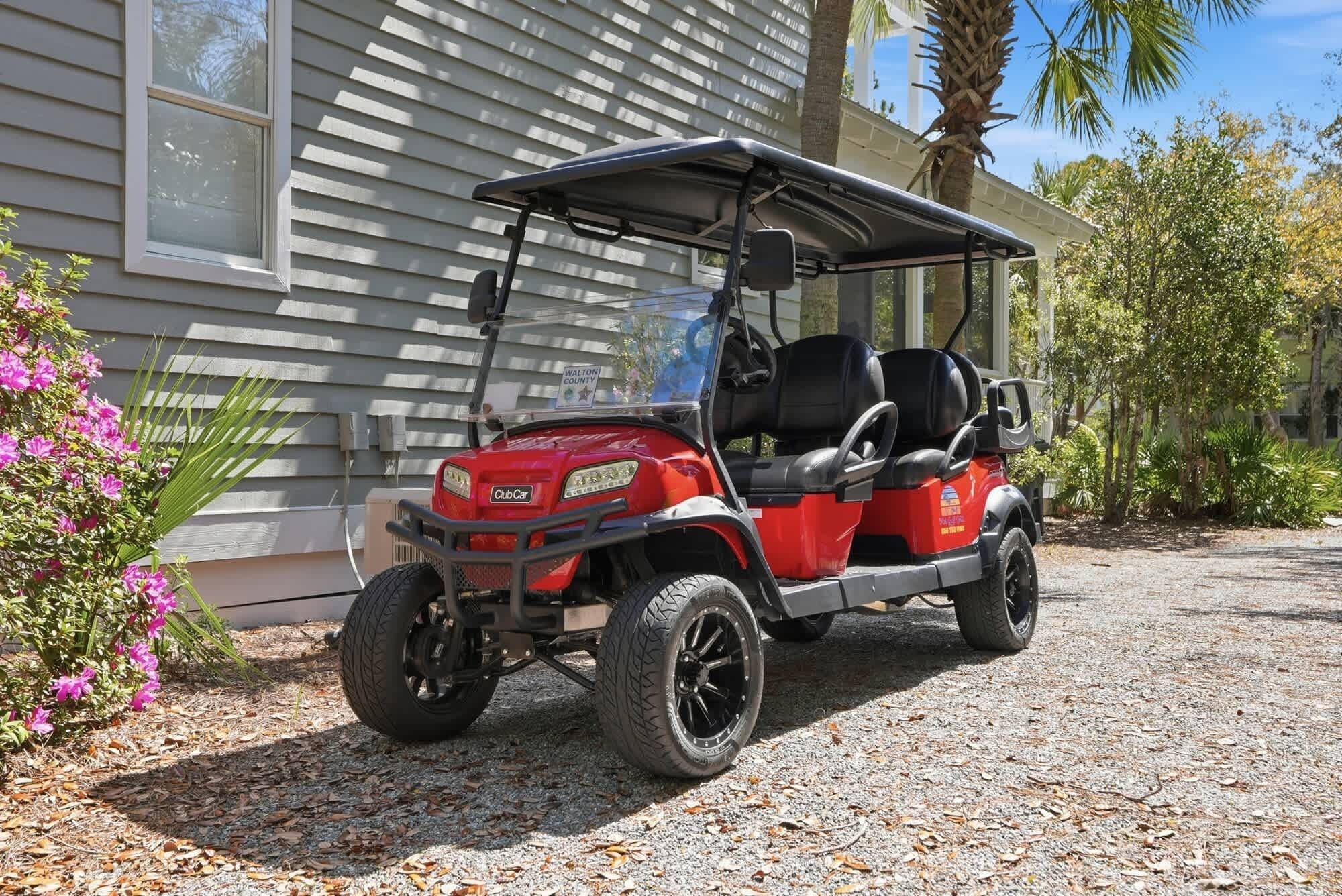 A red golf cart parked on a gravel driveway in front of a house with a white exterior and a palm tree nearby.