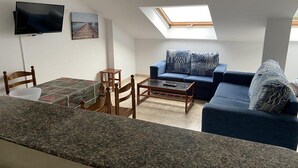 A modern living room with a blue sofa, a coffee table, a TV mounted on the wall, and a skylight. - Langosteira Apartaments (Fisterra)