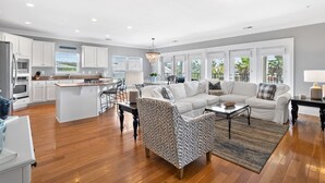 A spacious living area with a large sofa, a patterned armchair, and a coffee table. A kitchen with white cabinets and a dining area with a table and chairs are visible.