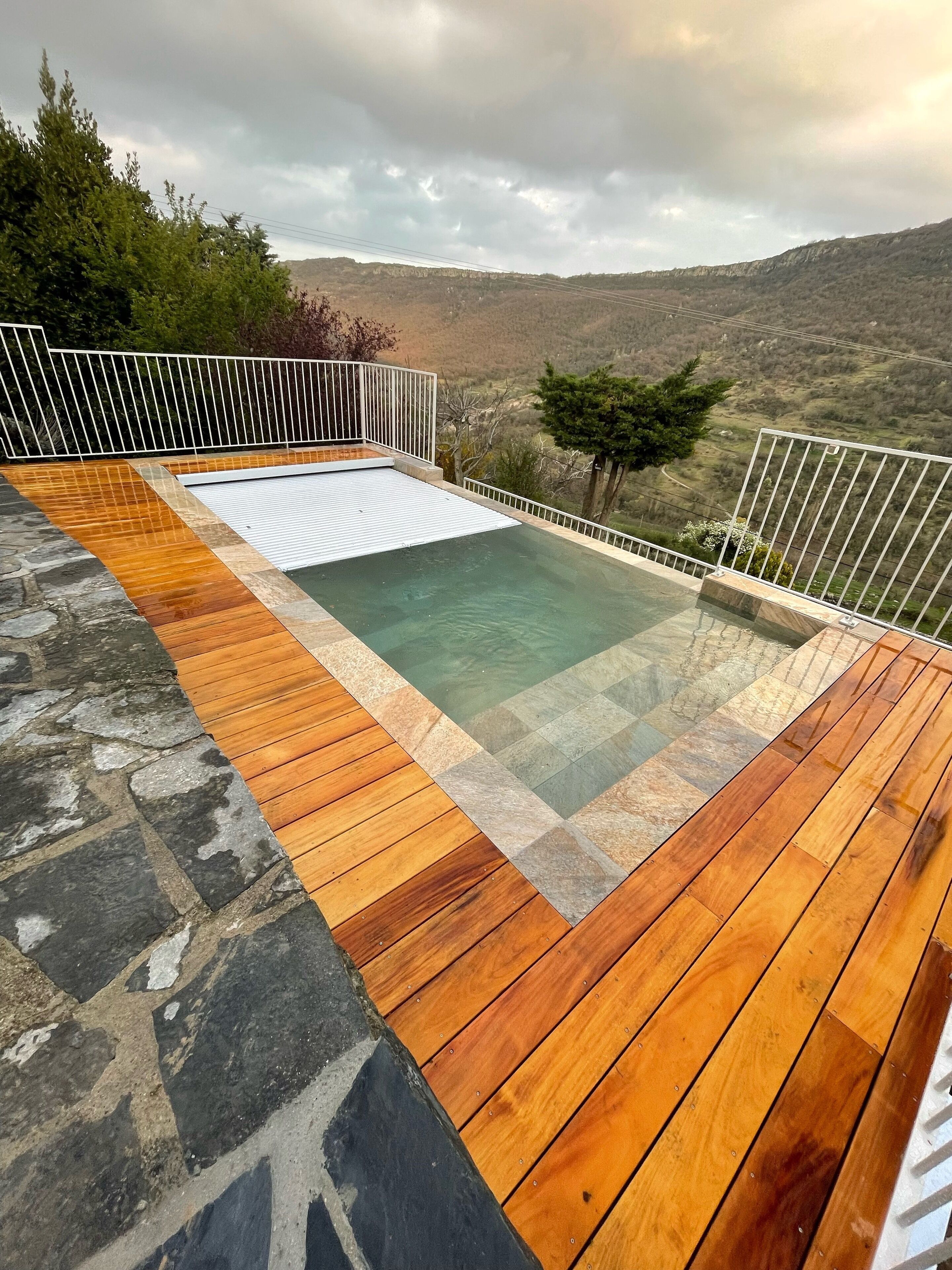 Una piscina infinita en la azotea con cubierta de madera y detalles de piedra, rodeada por una barandilla blanca, con vistas a un paisaje montañoso.