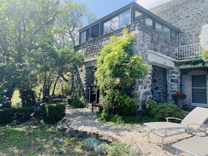 Une maison en pierre avec un balcon, un jardin agrémenté d’un chemin de gravier et un espace extérieur aménagé pour s’asseoir.
