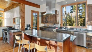 A modern kitchen with a large island, bar stools, and a view of trees through large windows. - Canyon View Retreat - Scenic & Secluded (Telluride)
