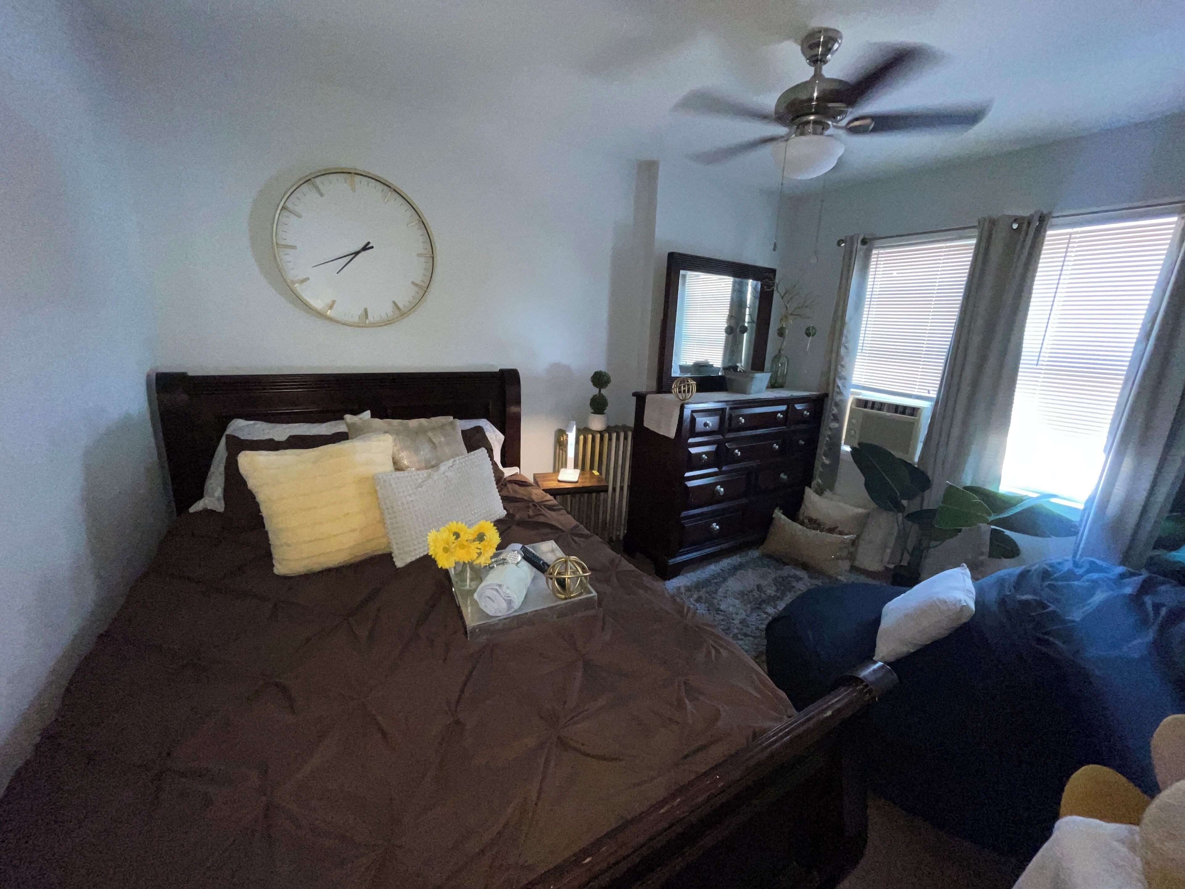 Une chambre à coucher comprenant un lit, une commode, une horloge, un ventilateur de plafond et une fenêtre avec des stores.