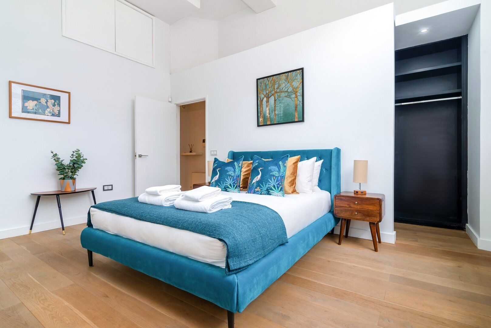 Een moderne slaapkamer met een groot bed, een nachtkastje, een plant in pot en een schilderij aan de muur.