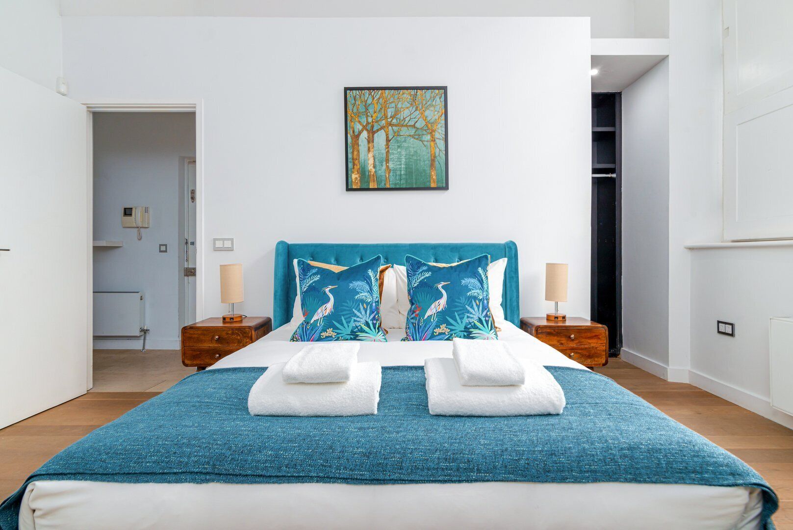 A modern bedroom with a large bed, two nightstands, a painting on the wall, and a view of the hallway.