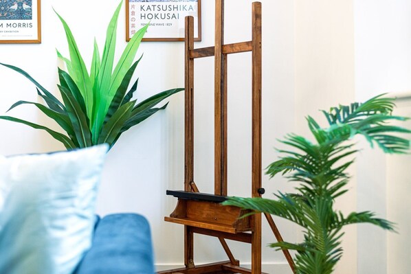 A wooden easel with a canvas, a potted plant, and a framed picture on a white wall.