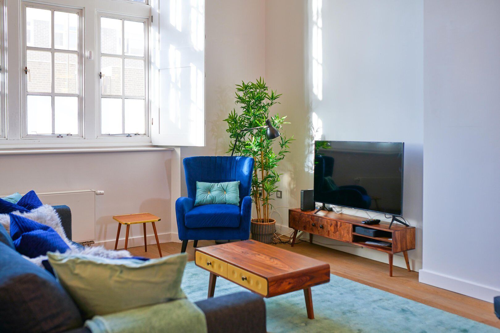A living room with a blue armchair, a wooden coffee table, a TV on a wooden stand, and a large window with white frames.