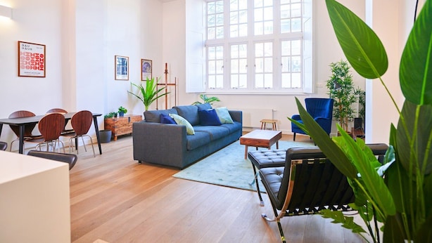 Uma sala de estar moderna com sofá, mesa de centro e cadeiras. Há janelas amplas, um espaço para refeições e diversas plantas.