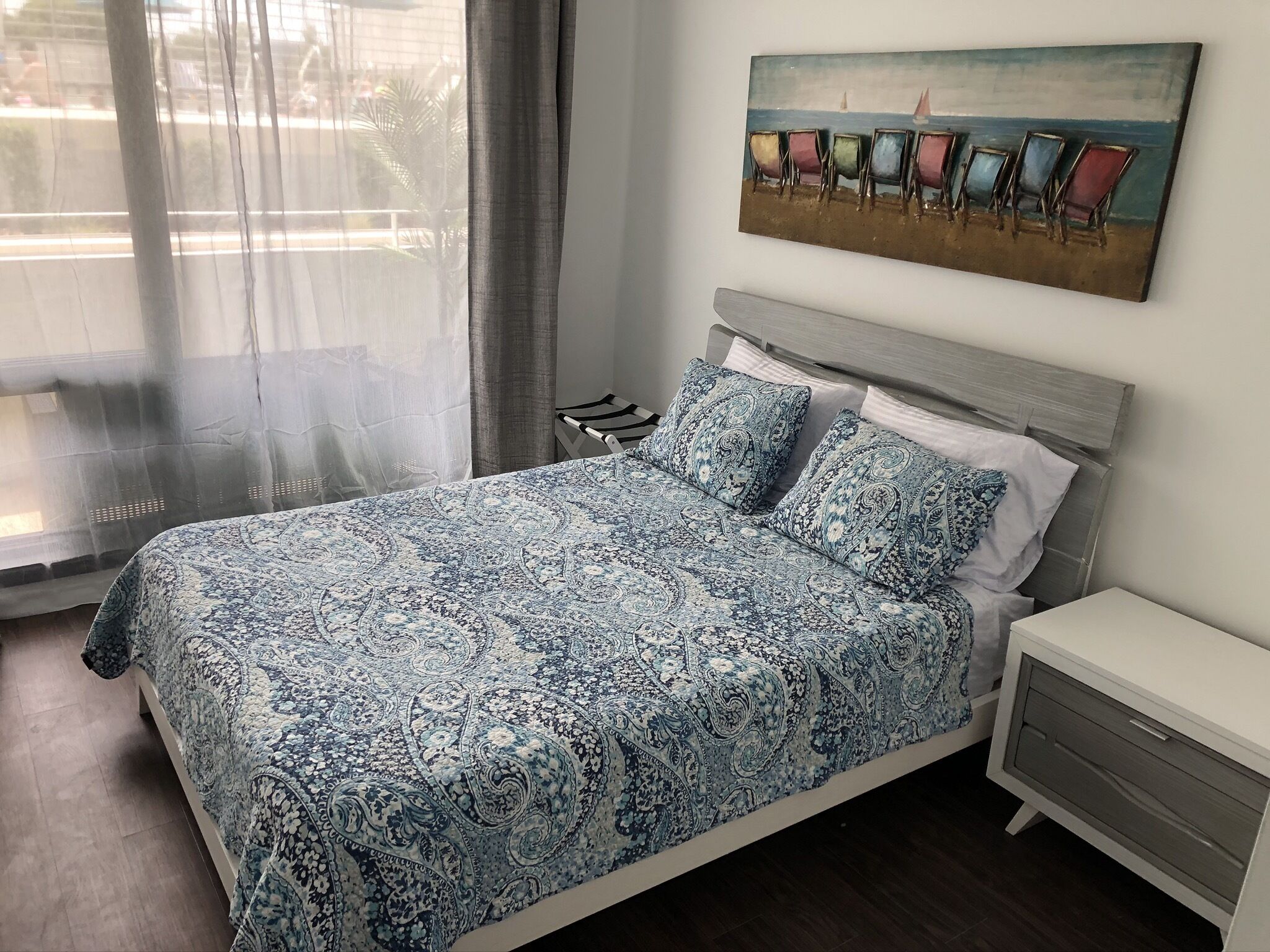 A bedroom with a bed, a nightstand, a painting, and a window with curtains.