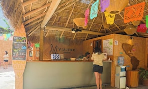 Un espace extérieur abrité par un toit de chaume, comprenant un comptoir, un distributeur d’eau et une enseigne « VIAJERO SAYULITA », ainsi qu’un panneau affichant le menu.