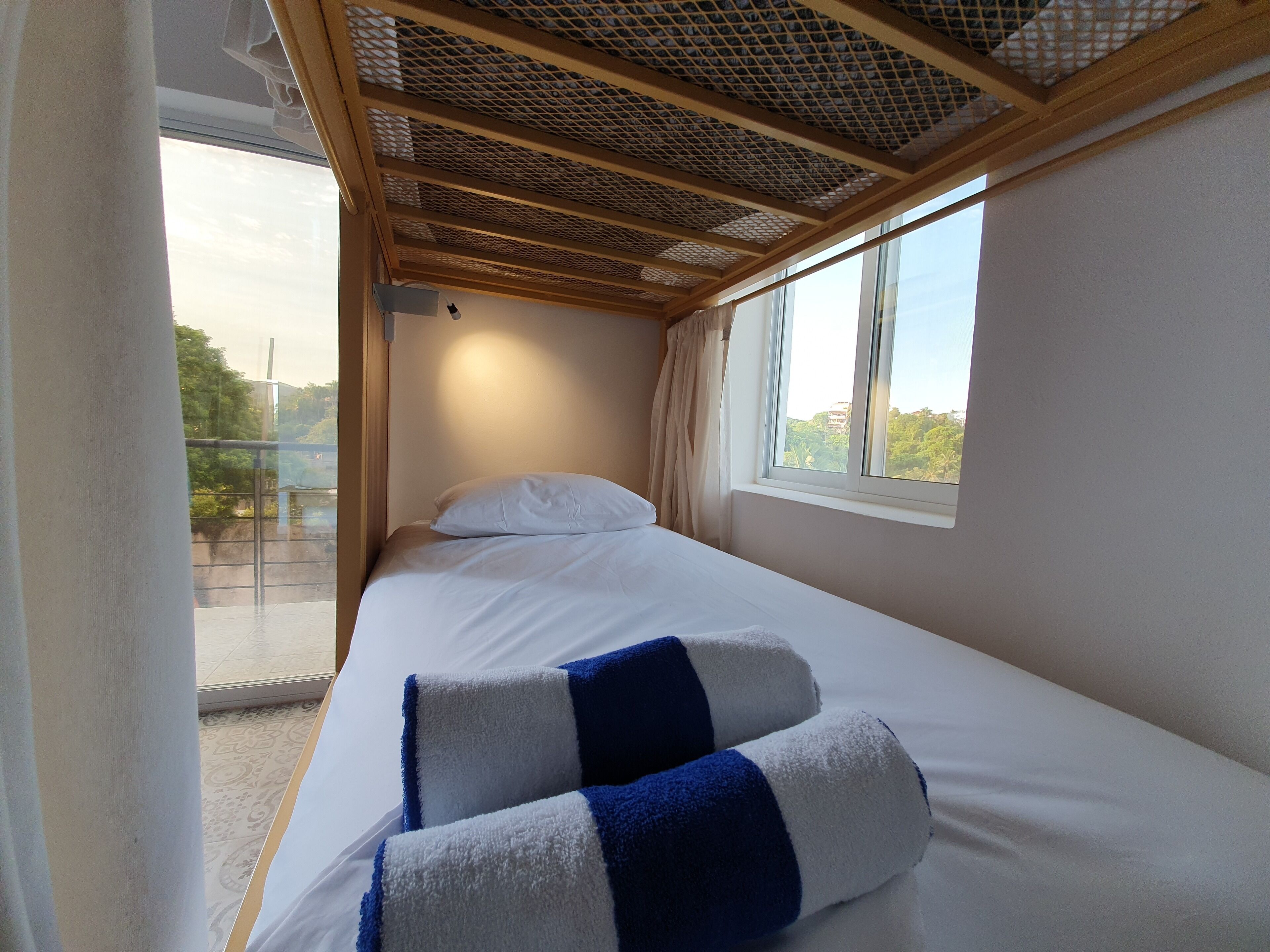 A single bed with white bedding and blue and white striped pillows, a window with a view of greenery, and a ceiling with a wooden lattice design.