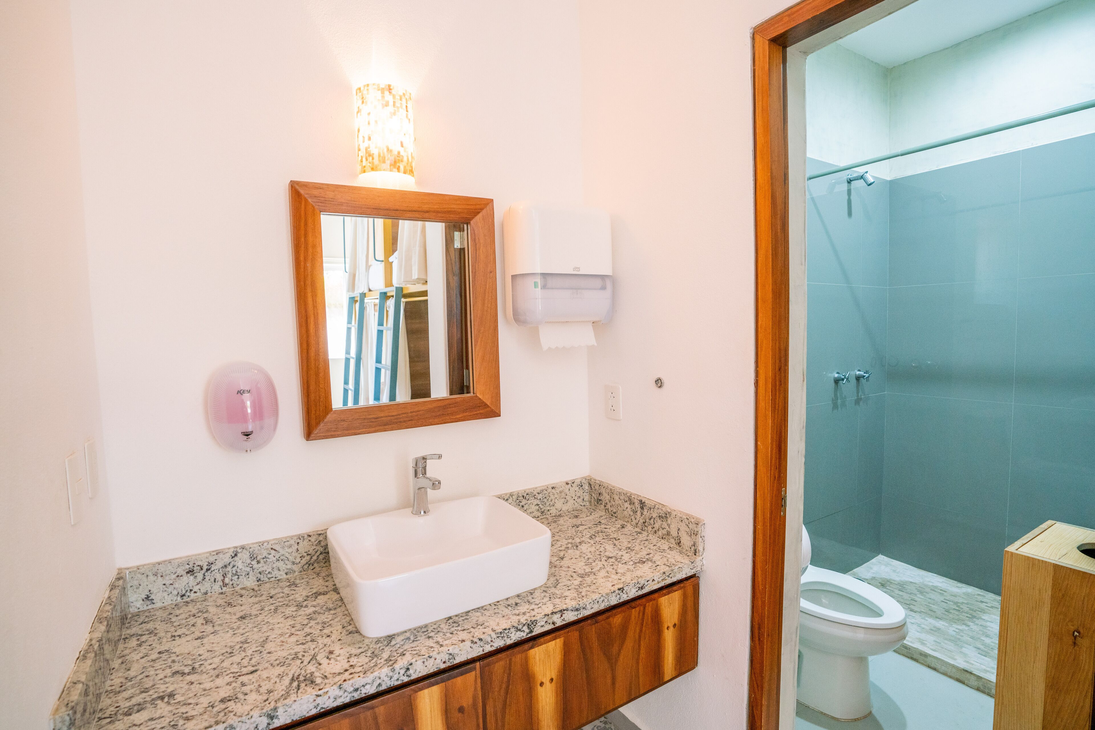 Un baño con encimera de mármol, tocador de madera, lavabo blanco, espejo, dispensador de jabón, dispensador de toallas de papel y cabina de ducha con paredes de vidrio.