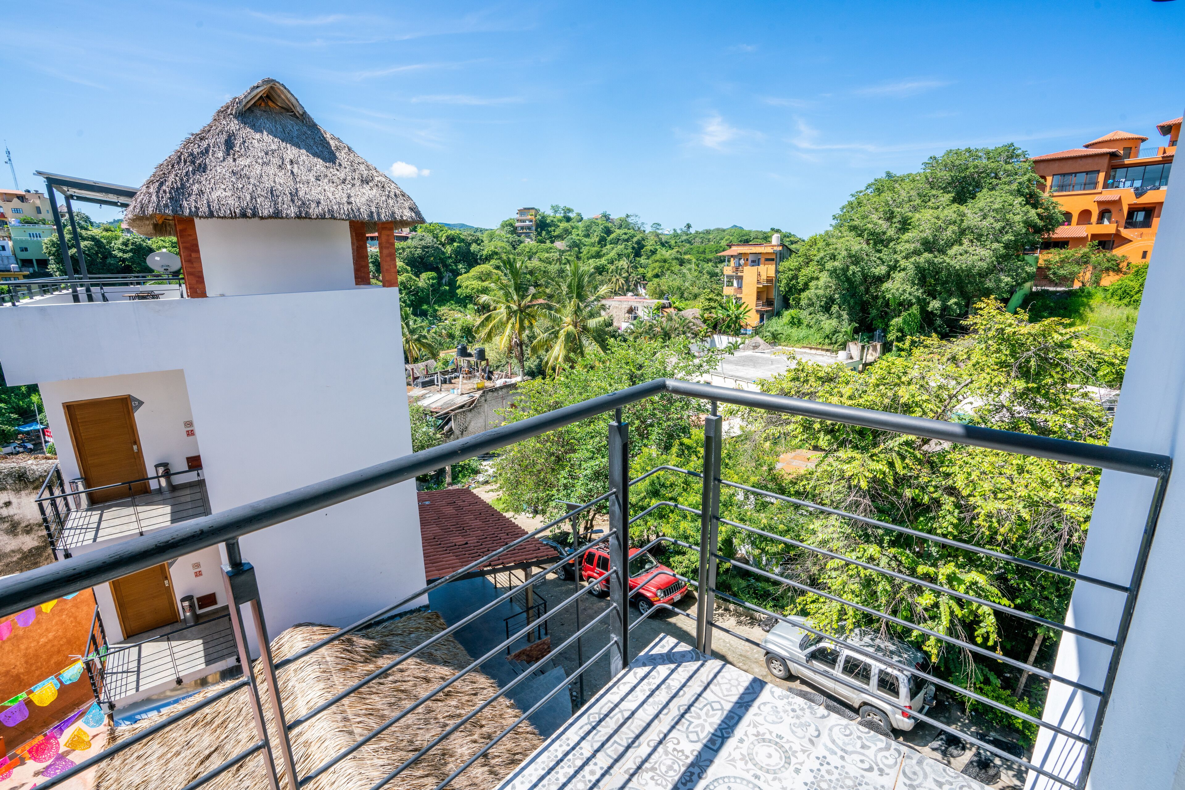 A balcony with a view of a red car, a thatched-roof structure, and a white building with a patterned floor.