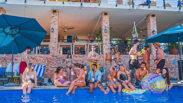 Un área junto a la piscina con gente sentada y de pie, una pelota inflable grande y sombrillas que dan sombra.