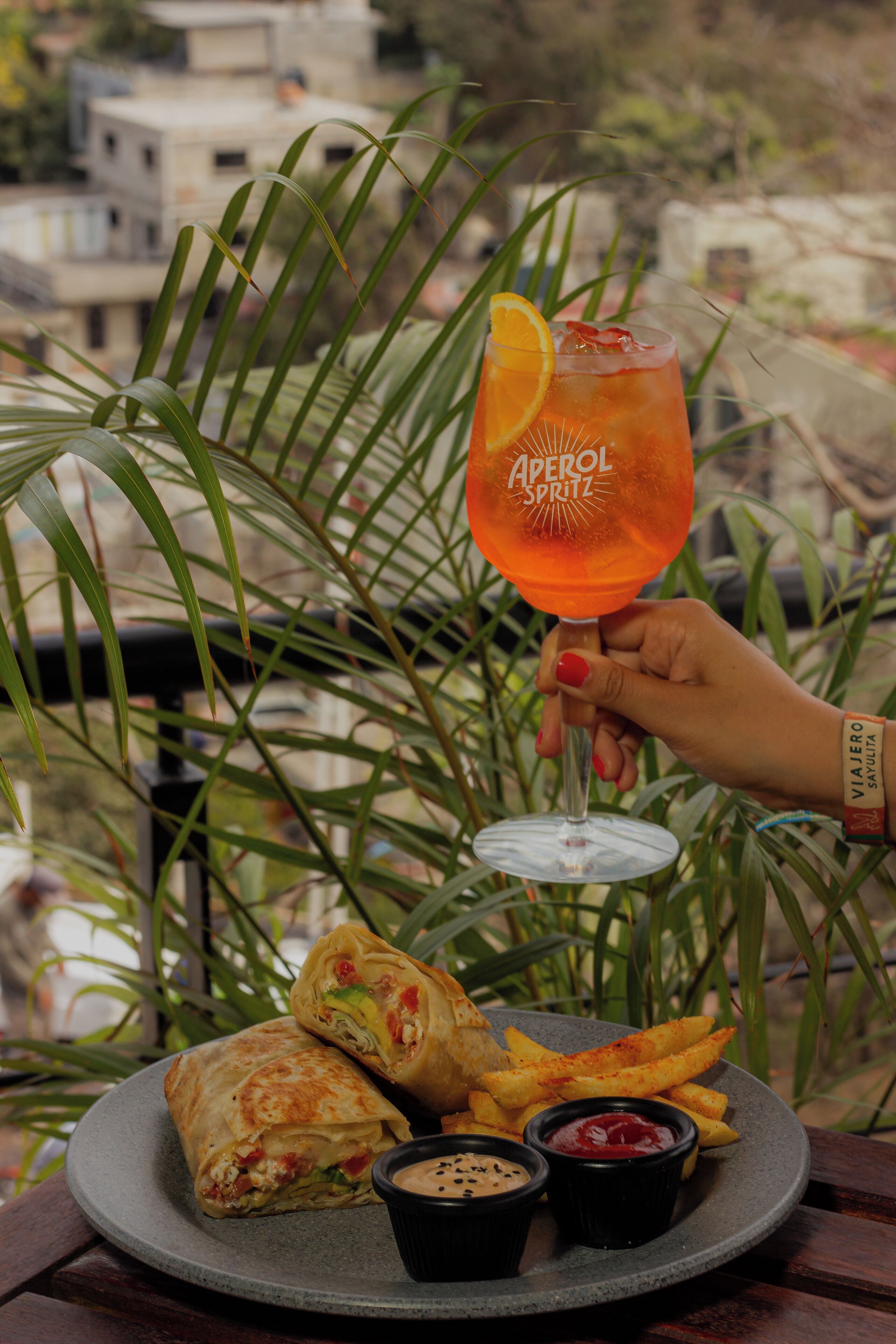 Une assiette comprenant un sandwich enroulé dans une tortilla, des frites et deux sauces pour tremper, accompagnée d'un verre d'Aperol Spritz.