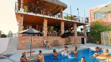 Un área de piscina con una estructura de techo de paja, asientos al aire libre y un edificio de ladrillo.