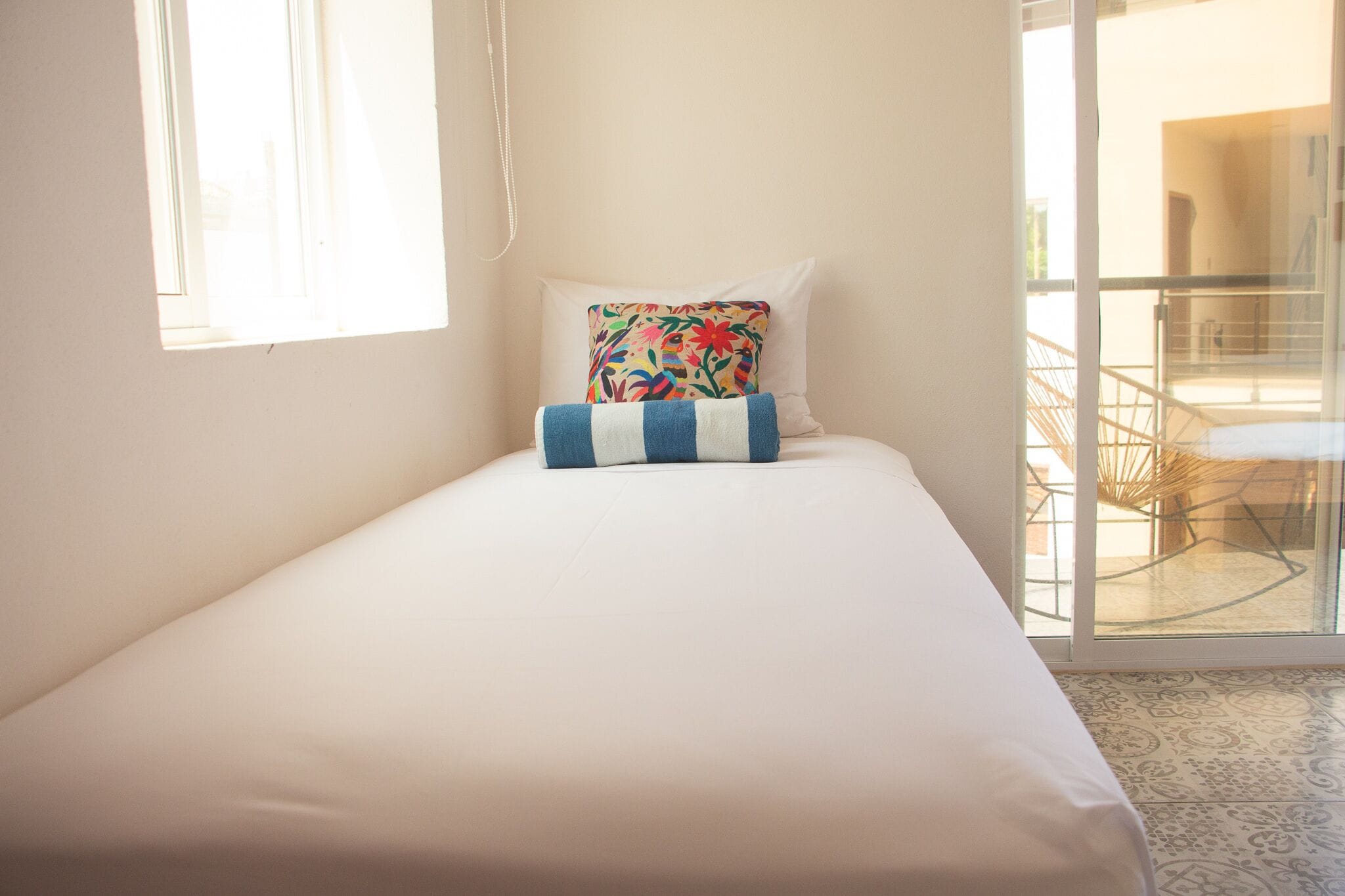 Un lit simple avec une literie blanche et un coussin à motifs floraux colorés, une fenêtre avec des stores et une vue sur un balcon extérieur.