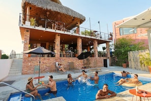 Un espace piscine comprenant une structure à toit de chaume, un bâtiment en briques et un bâtiment moderne doté d’un balcon.