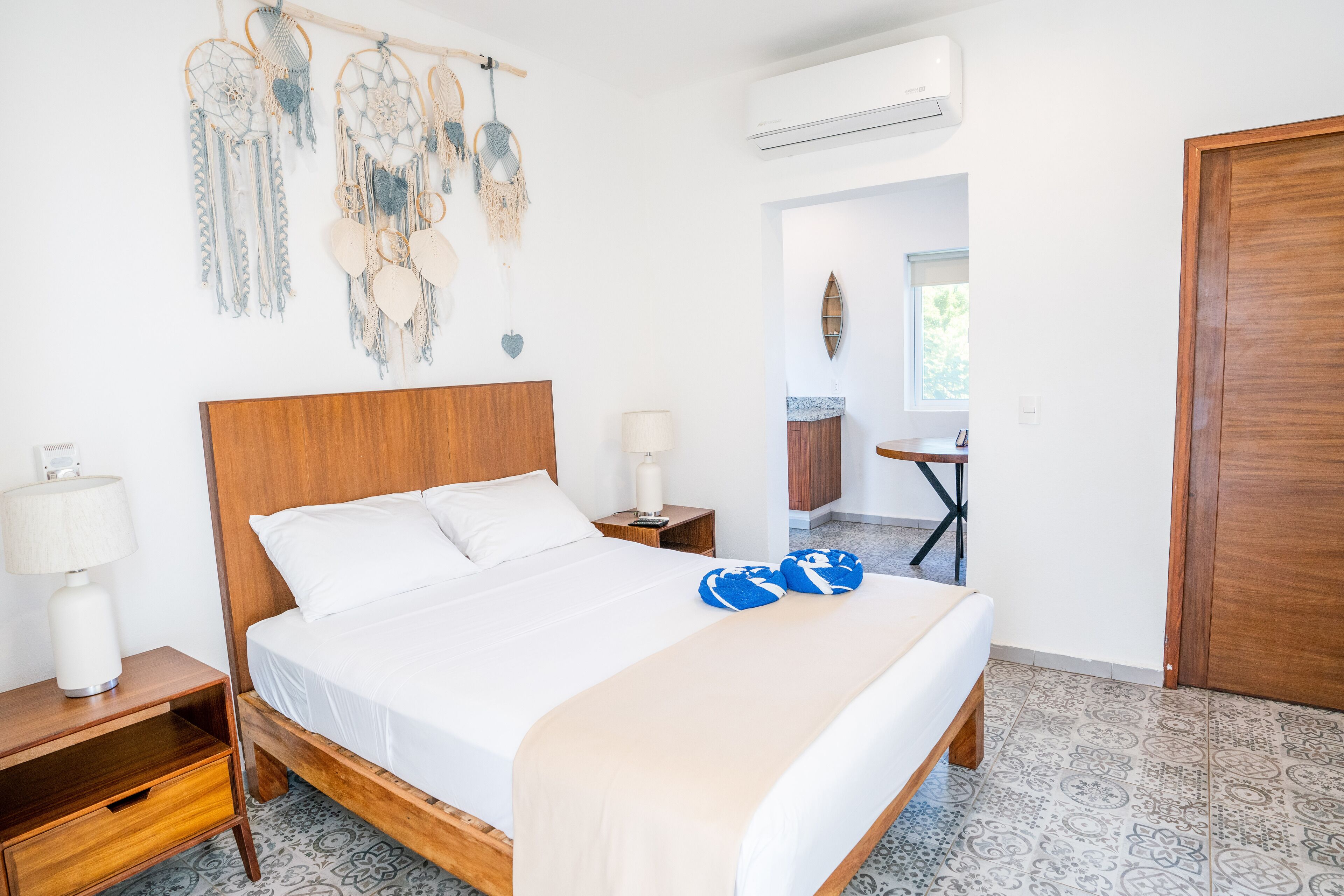 A neatly made bed with a wooden headboard, a bedside table with a lamp, and a wall decorated with macrame dreamcatchers.