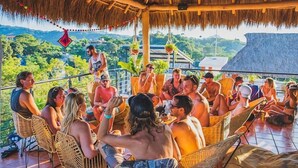 Un grupo de personas sentadas en sillas de mimbre bajo un techo de paja, con vistas a un paisaje exuberante.